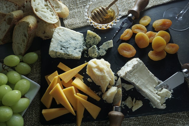 Slices of assorted cheese with fresh and dried fruits, honey, and slices of bread on a black board