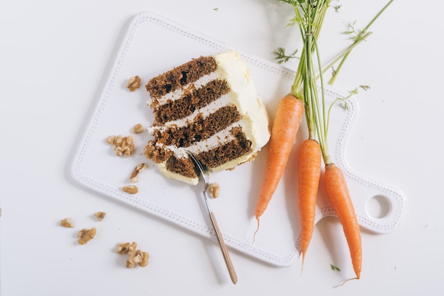 A slice of carrot cake on a white chopping board with pieces of carrots on the side