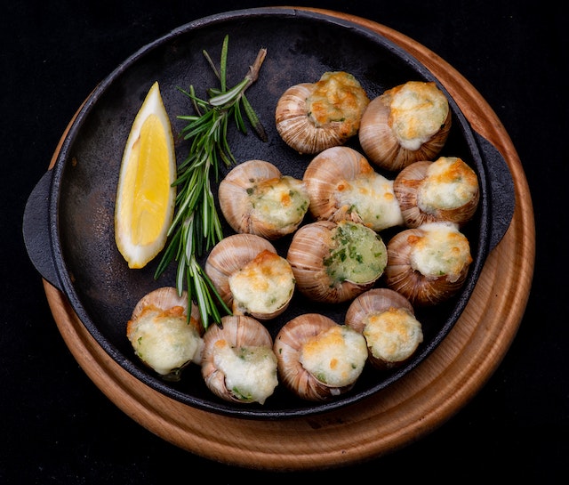 Cooked escargot in a black baking dish, served with a slice of lemon and some rosemary